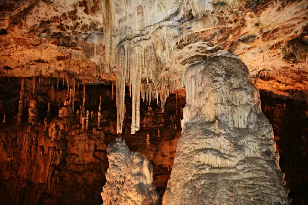 Stalaktity a stalagmity v jeskyni Moravského krasu, osvětlené teplým světlem, které zvýrazňuje detaily těchto přírodních útvarů. Na stropě visí dlouhé stalaktity, zatímco ze země se tyčí mohutné stalagmity, vytvářejíce fascinující podzemní scenérii.