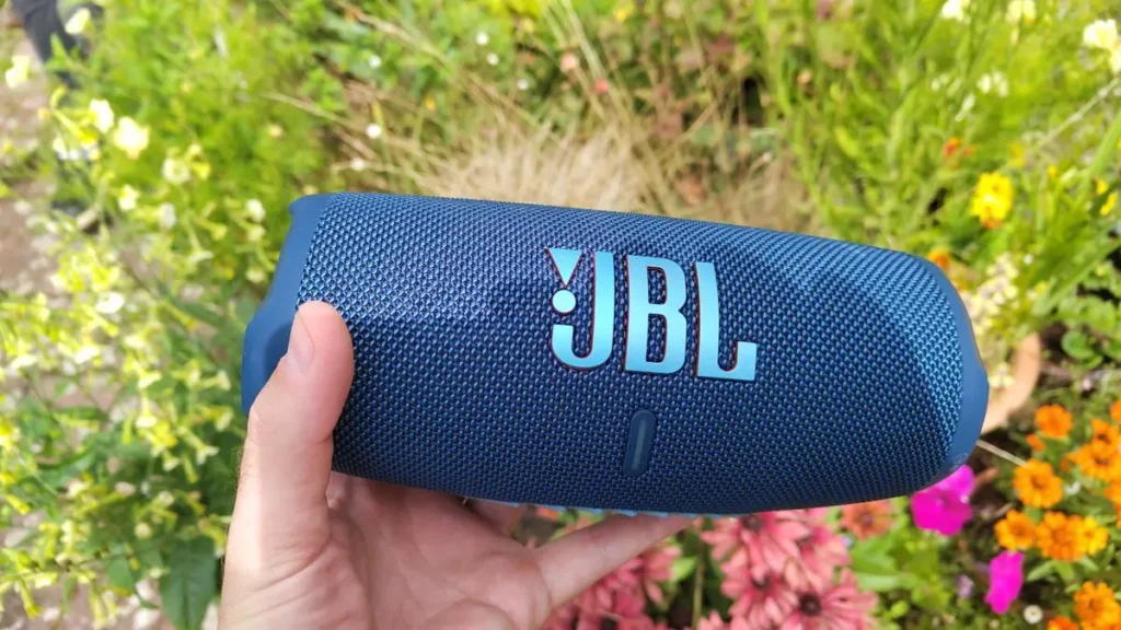 JBL Charge 5 bluetooth reproduktor v modré barvě držený v ruce s květinovým pozadím na zahradě.