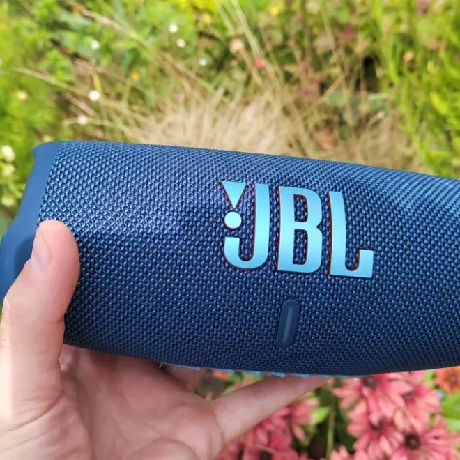 JBL Charge 5 bluetooth reproduktor v modré barvě držený v ruce s květinovým pozadím na zahradě.