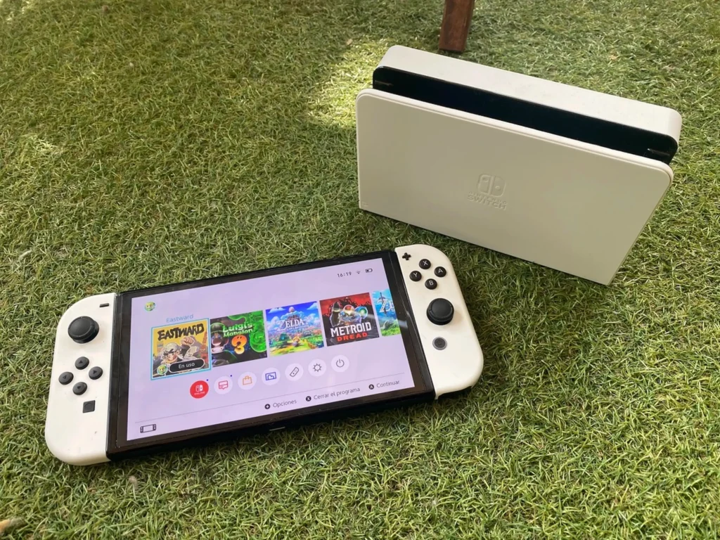 Herní konzole Nintendo Switch OLED s bílými Joy-Con ovladači a dokovací stanicí na trávníku.