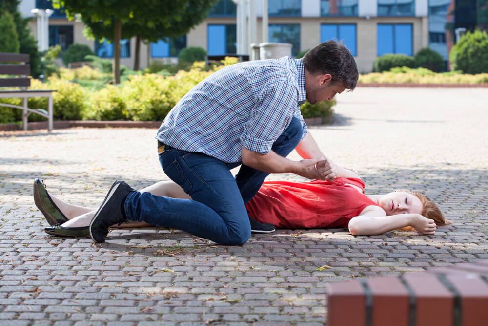 Muž poskytuje první pomoc ženě ležící na zemi, která má zřejmě epileptický záchvat. Žena je položena na bok na ulici v parku, zatímco muž se jí snaží pomoci a zajistit její bezpečnost.