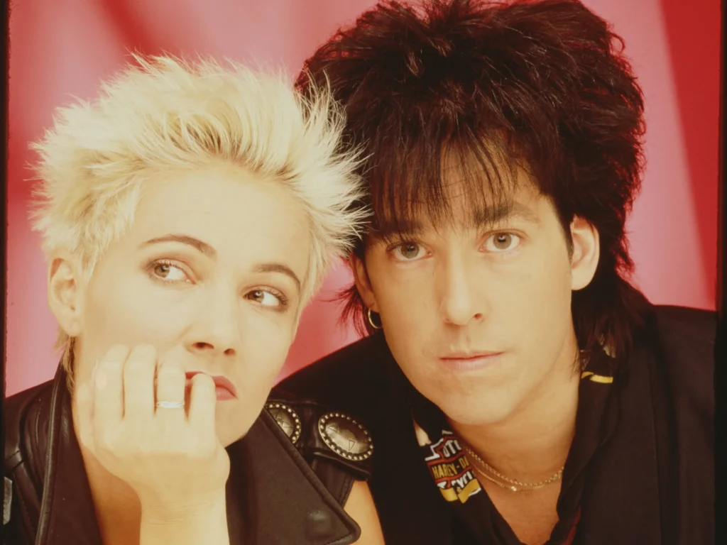 Marie Fredriksson a Per Gessle z Roxette v 80. letech, pózující před růžovým pozadím. Marie má zamyšlený výraz, zatímco Per se dívá přímo do kamery.