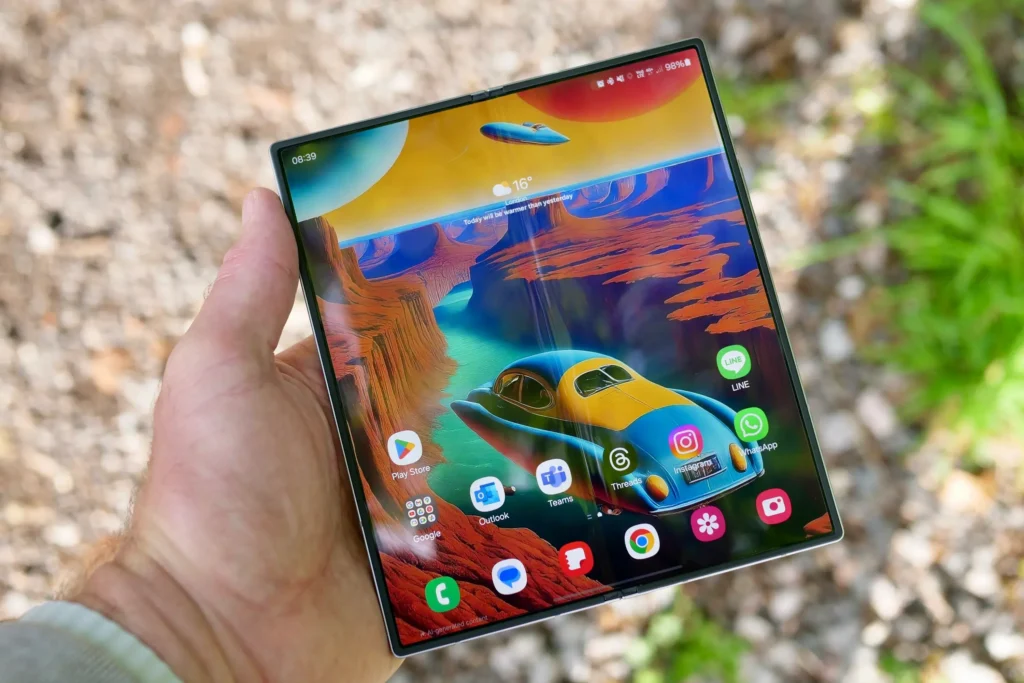 Ruka držící rozložený mobilní telefon Samsung Galaxy Z Fold6 s barevným a jasným AMOLED displejem, na kterém jsou zobrazeny různé aplikace. Telefon má futuristický design s velkou obrazovkou, ideální pro multimediální zábavu a multitasking.