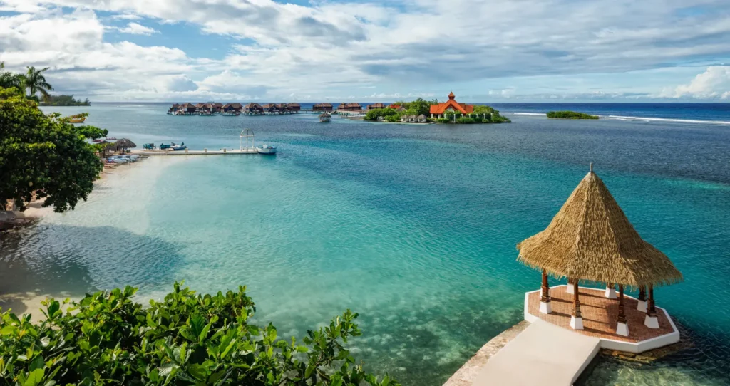 Pohled na klidnou lagunu a soukromý ostrov v resortu Sandals Royal Caribbean na Jamajce, s malým altánkem s doškovou střechou, kde si můžete vychutnat romantické chvíle s výhledem na moře.