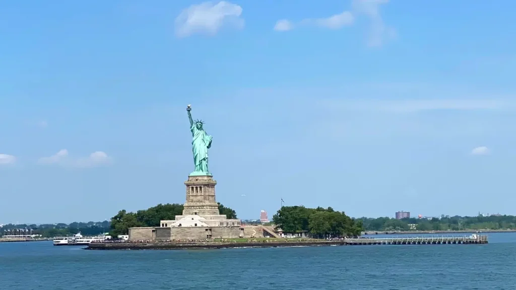 Socha Svobody stojící na Liberty Islandu v New Yorku za jasného dne, s výhledem na přilehlé vody a oblohu.