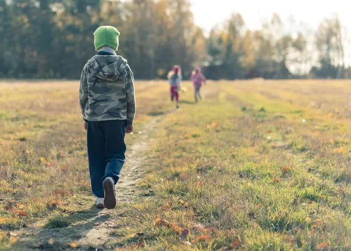 Na obrázku je dítě v zelené čepici a maskáčové bundě, které kráčí po úzké pěšině vedoucí travnatou loukou. V pozadí jsou rozmazaně vidět další děti, které běží směrem od kamery. Scéna se odehrává za podzimního počasí, kdy je okolí pokryté spadaným listím a v dálce jsou vidět stromy.