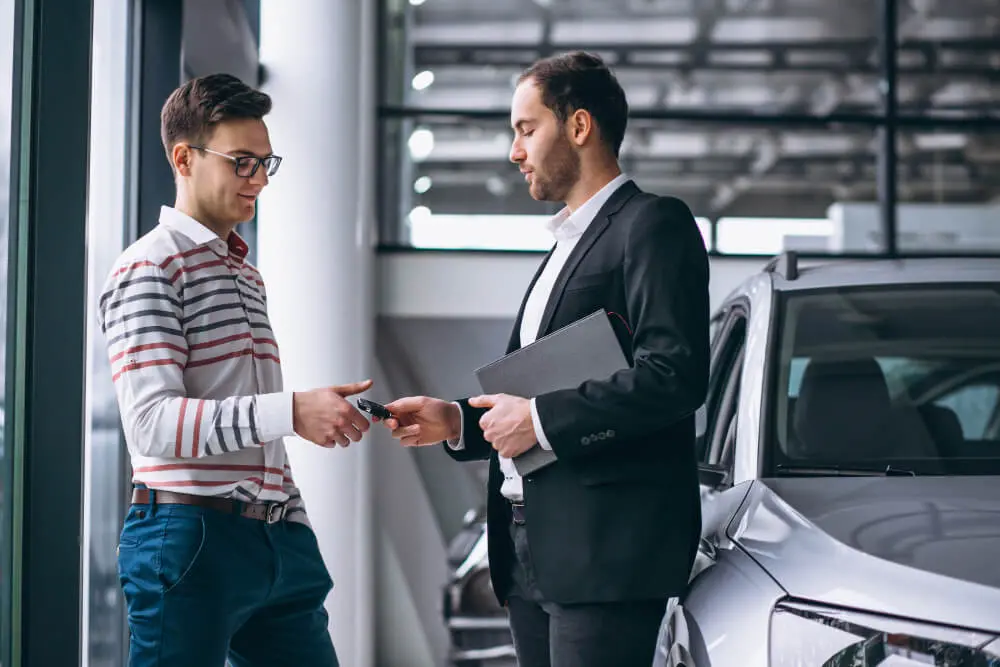 Muž v obleku předává klíče od auta druhému muži ve světlém svetru uvnitř autosalonu. Oba muži se nacházejí vedle stříbrného vozu. Scéna symbolizuje uzavření kupní smlouvy na auto.