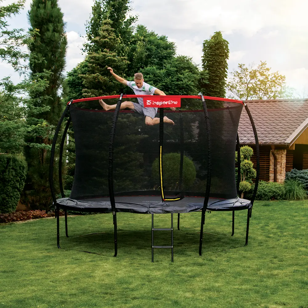 Muž skáče na stojící trampolíně značky inSPORTline s ochrannou sítí, umístěné na zelené zahradě před domem.