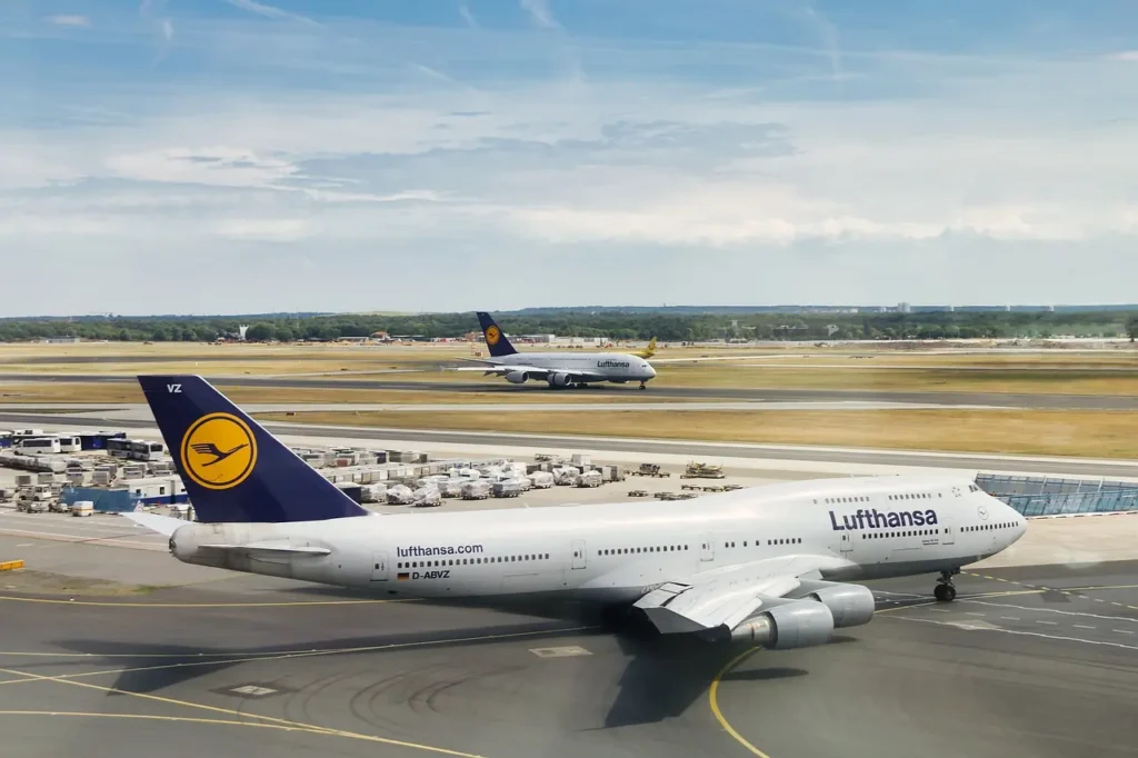 Boeing 747 letecké společnosti Lufthansa na letišti, připravující se na vzlet, s dalším letadlem stejné společnosti v pozadí na přistávací dráze.