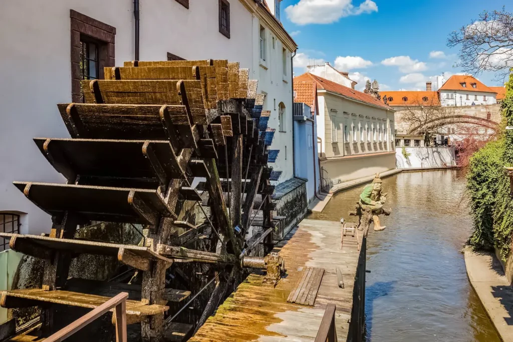 Historické mlýnské kolo na kanálu Čertovka v Praze, obklopené malebnou architekturou a klidnou vodní hladinou za slunečného dne.