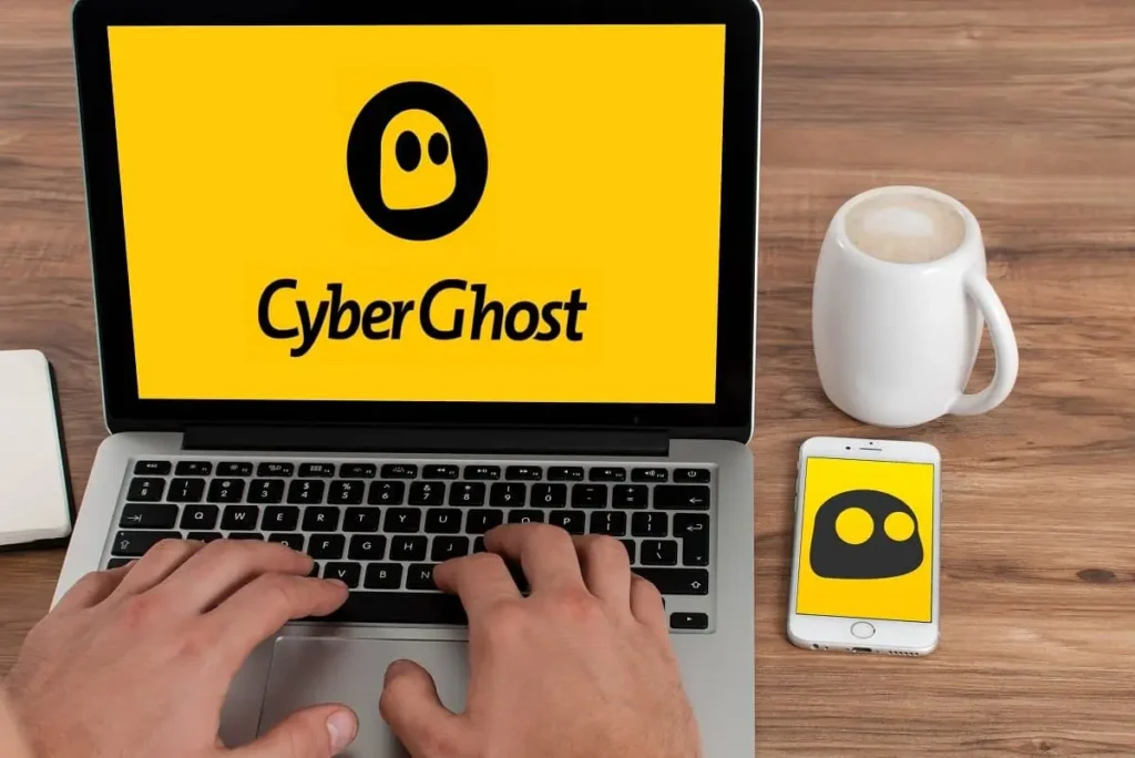 Uživatel pracuje na laptopu, který zobrazuje logo CyberGhost VPN, zatímco vedle je umístěný telefon s aplikací CyberGhost a šálkem kávy.