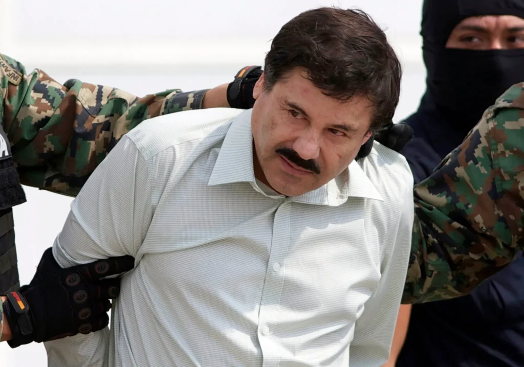 Joaquín 'El Chapo' Guzmán v bílém košili, zadržený mexickými bezpečnostními složkami. Obrázek zachycuje moment po jeho zatčení, známý drogový boss je veden do vazby