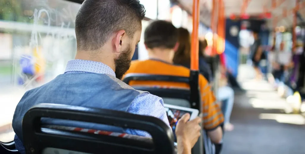 Cestující v tramvaji používající tablet připojený k bezplatné Wi-Fi, zachycen zezadu v běžné situaci během přepravy.