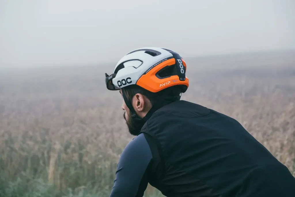 Muž na kole se dívá do dálky v mlhavé krajině, na hlavě má cyklistickou přilbu značky POC s oranžovo-bílým designem.