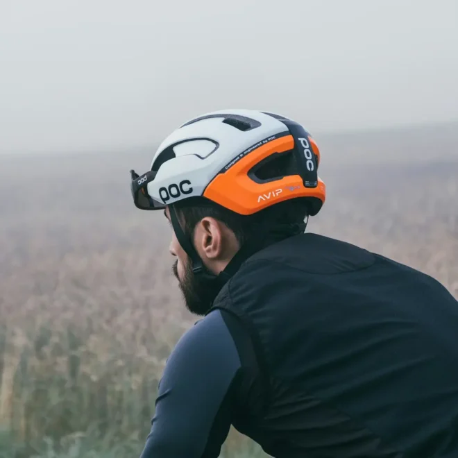 Muž na kole se dívá do dálky v mlhavé krajině, na hlavě má cyklistickou přilbu značky POC s oranžovo-bílým designem.