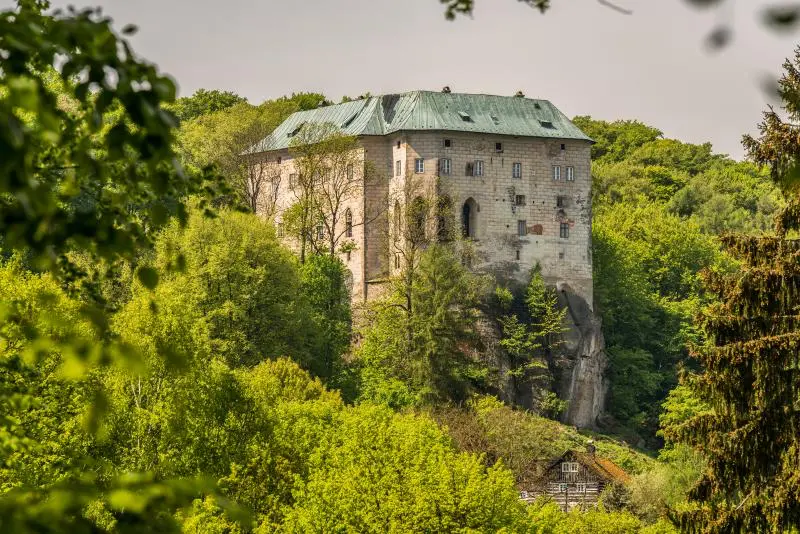 Středověký hrad Houska obklopený hustou zelení, nacházející se na vrcholu skalního masivu. Hrad je známý svou záhadnou historií a pověstmi o portálu do jiného světa.