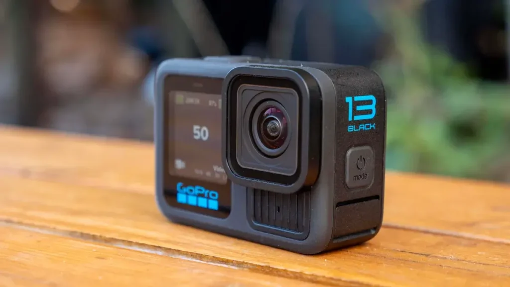 GoPro Hero 13 Black - Přední pohled na kameru: Pohled zepředu na GoPro Hero 13 Black, zobrazující přední objektiv a malé displeje kamery na dřevěném stole.