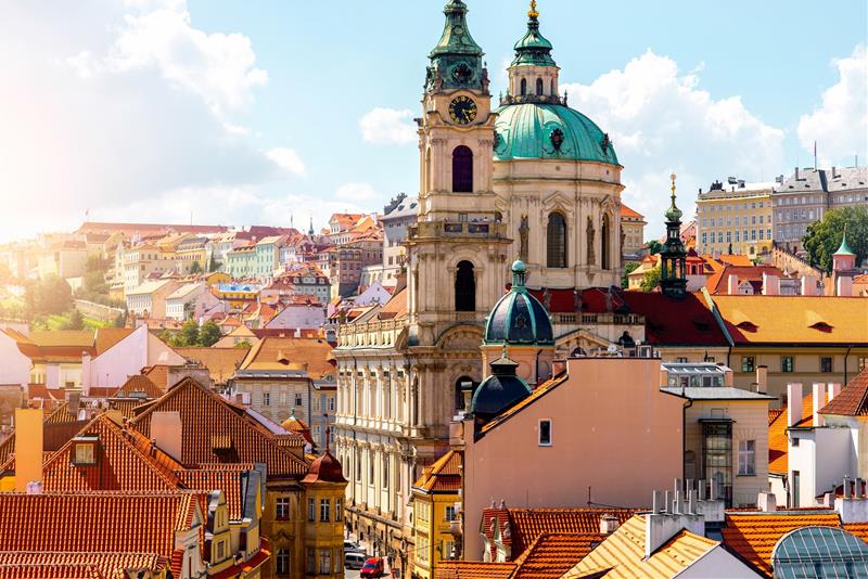 Kostel svatého Mikuláše na Malé Straně v Praze, obklopený historickými budovami a osvětlený slunečním světlem za jasného dne.