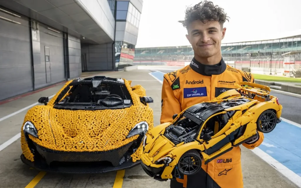 Lando Norris drží model LEGO® Technic McLaren P1™ vedle plně sestaveného skutečného modelu McLaren P1 z LEGO kostek na závodní dráze.