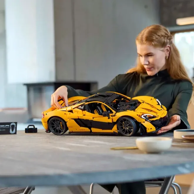 Žena sestavuje detailní model LEGO® Technic McLaren P1™ u stolu, soustředí se na pohyblivé části modelu.