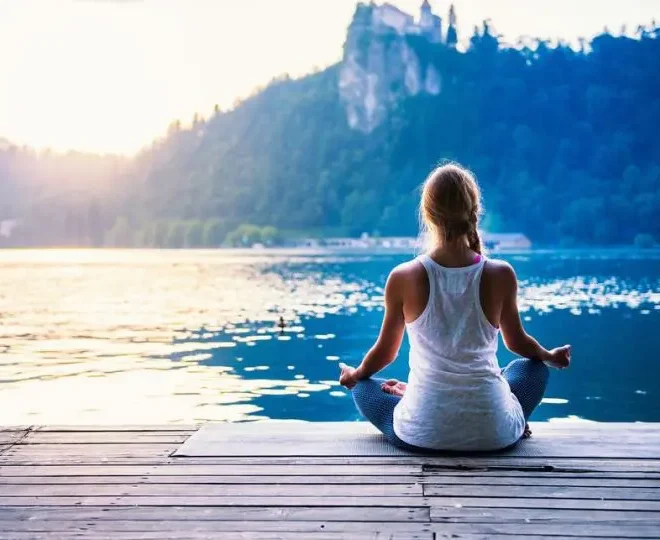 Žena sedí v meditační pozici na dřevěném molu u jezera s výhledem na hory při západu slunce, obklopena klidnou přírodou.