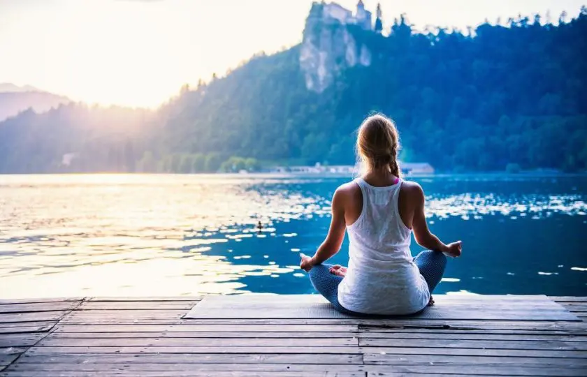 Žena sedí v meditační pozici na dřevěném molu u jezera s výhledem na hory při západu slunce, obklopena klidnou přírodou.