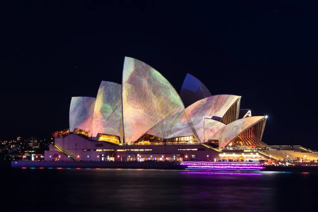 Sydney Opera House během noční světelné show – slavná budova opery v Sydney je osvětlena barevnými světelnými projekcemi, které vytvářejí poutavé vizuální efekty na jejích výrazných střechách. Noční atmosféru doplňuje odraz světel na hladině vody v přístavu.