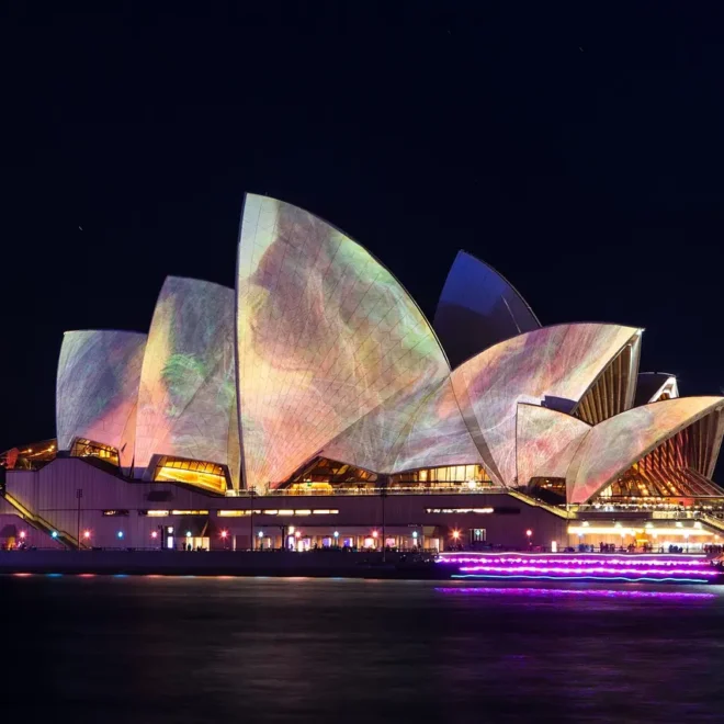 Sydney Opera House během noční světelné show – slavná budova opery v Sydney je osvětlena barevnými světelnými projekcemi, které vytvářejí poutavé vizuální efekty na jejích výrazných střechách. Noční atmosféru doplňuje odraz světel na hladině vody v přístavu.