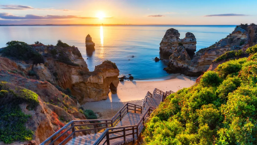 Západ slunce nad malebnou pláží v Algarve, Portugalsko, s dřevěnými schody vedoucími na písečnou pláž mezi dramatickými útesy a klidným, průzračným mořem.