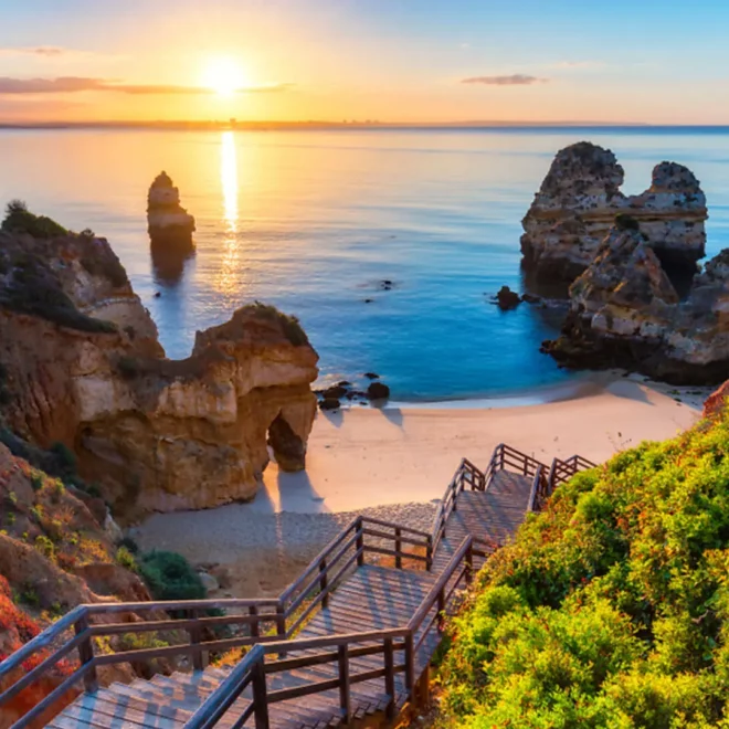 Západ slunce nad malebnou pláží v Algarve, Portugalsko, s dřevěnými schody vedoucími na písečnou pláž mezi dramatickými útesy a klidným, průzračným mořem.