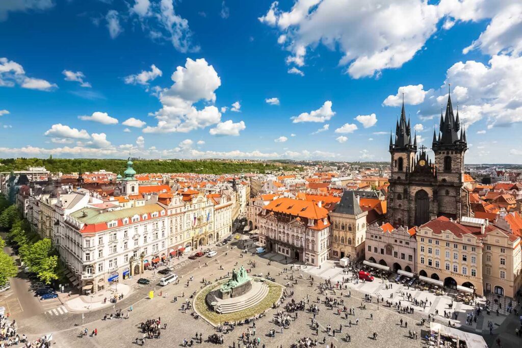 Letecký pohled na historické centrum Prahy, Staroměstské náměstí s Týnským chrámem a okolními budovami. Jasná obloha s bílými mraky a živým ruchem na náměstí dotváří atmosféru jednoho z nejznámějších pražských míst.