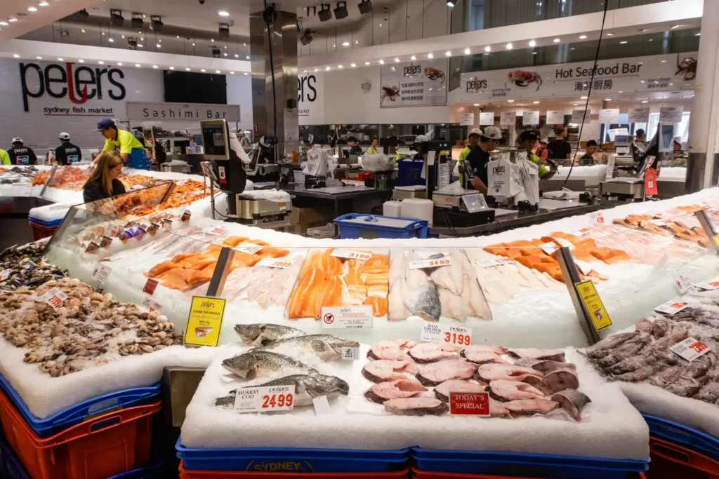 Sydney Fish Market – vnitřní pohled na trh s mořskými plody, kde jsou na ledu vystaveny různé druhy čerstvých ryb a mořských plodů. Zákazníci vybírají z široké nabídky, zatímco zaměstnanci pracují za pultem. Na trhu je vidět jasné značení cen a nabízených produktů.