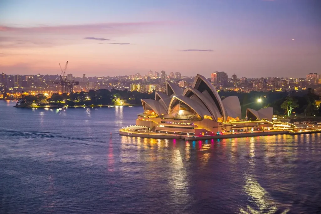 Sydney Opera House při západu slunce – ikonická budova opery v Sydney zářící v nočním osvětlení, odráží se ve vodách přístavu. V pozadí jsou osvětlené mrakodrapy města, vytvářející působivou scenérii