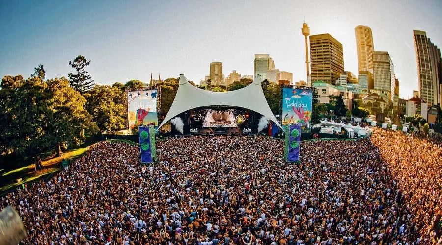 Hudební festival v Sydney – obrovský dav návštěvníků si užívá živou hudbu pod širým nebem na festivalu v Sydney. V popředí je přeplněné publikum před velkým pódiem s výrazným bílým stanem, zatímco v pozadí se tyčí moderní mrakodrapy města.