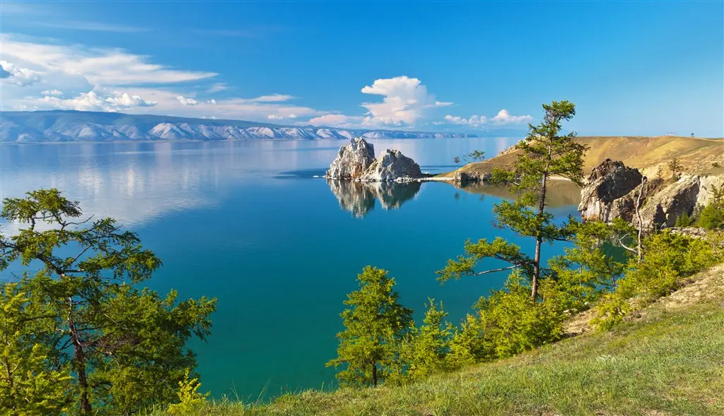 Malebný pohled na jezero obklopené zelenými kopci a skalnatými výběžky, s průzračnou vodou odrážející oblohu a vzdálené pohoří.