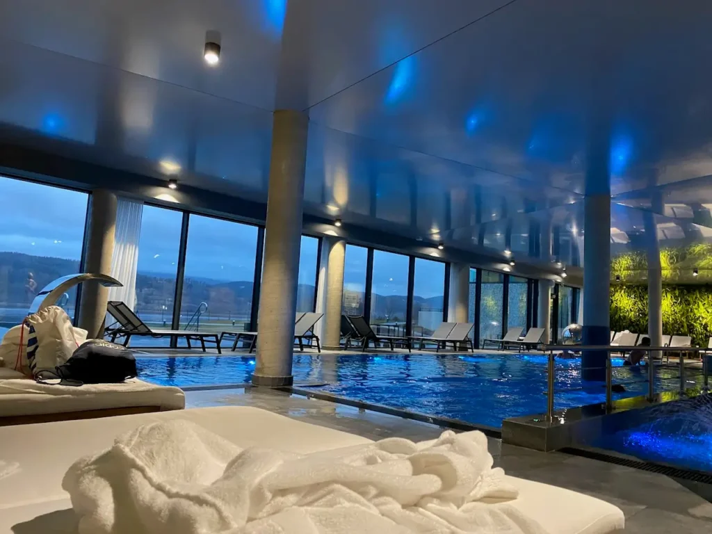 Moderní wellness zóna v Lake Hill Resort & Spa s bazénem a pohodlnými lehátky, výhled na jezero a okolní hory, modré osvětlení vytváří klidnou atmosféru.