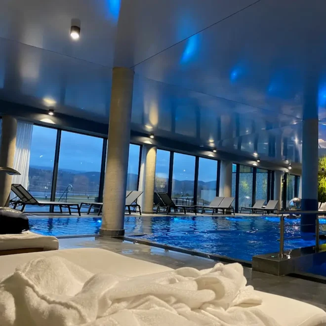 Moderní wellness zóna v Lake Hill Resort & Spa s bazénem a pohodlnými lehátky, výhled na jezero a okolní hory, modré osvětlení vytváří klidnou atmosféru.