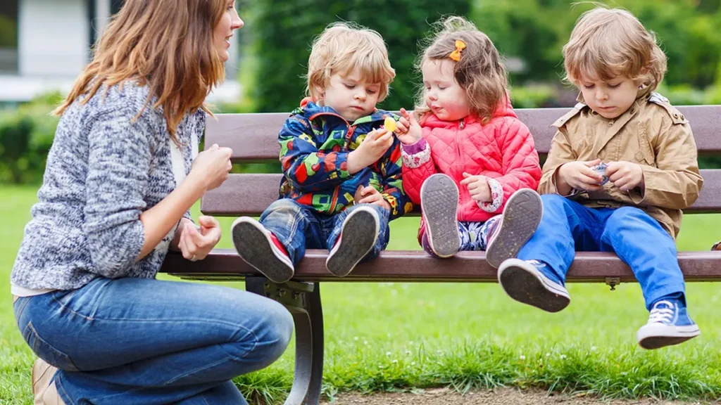 Matka sedící na lavičce v parku se třemi malými dětmi v barevných bundách, které si hrají s malými předměty. Děti jsou usazeny vedle sebe a matka je sleduje s úsměvem.