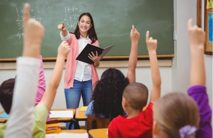 Učitelka vyvolává žáky ve třídě, zatímco děti s nadšením zvedají ruce. Na tabuli jsou jednoduché matematické úlohy. Atmosféra ve třídě je plná energie a nadšení z učení.