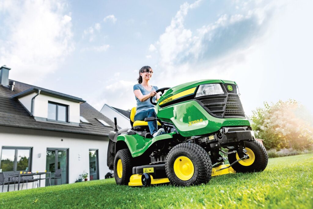 Žena řídící zahradní traktor John Deere X350R na udržovaném trávníku před moderním domem.