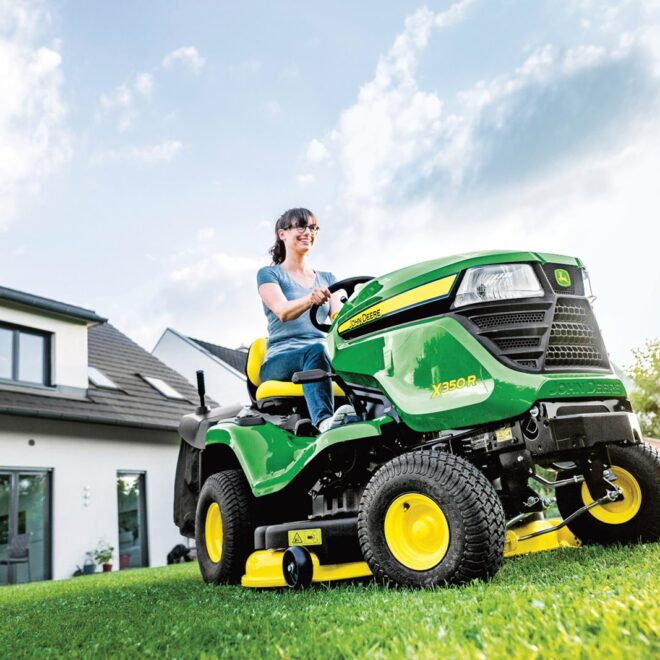 Žena řídící zahradní traktor John Deere X350R na udržovaném trávníku před moderním domem.