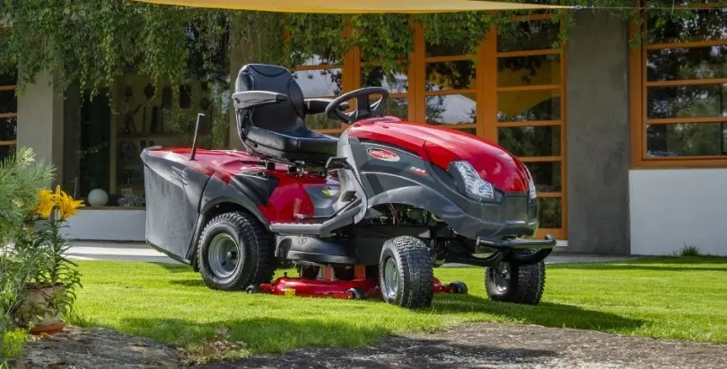 Červený zahradní traktor Mountfield stojící na udržovaném trávníku před moderním domem s velkými okny.