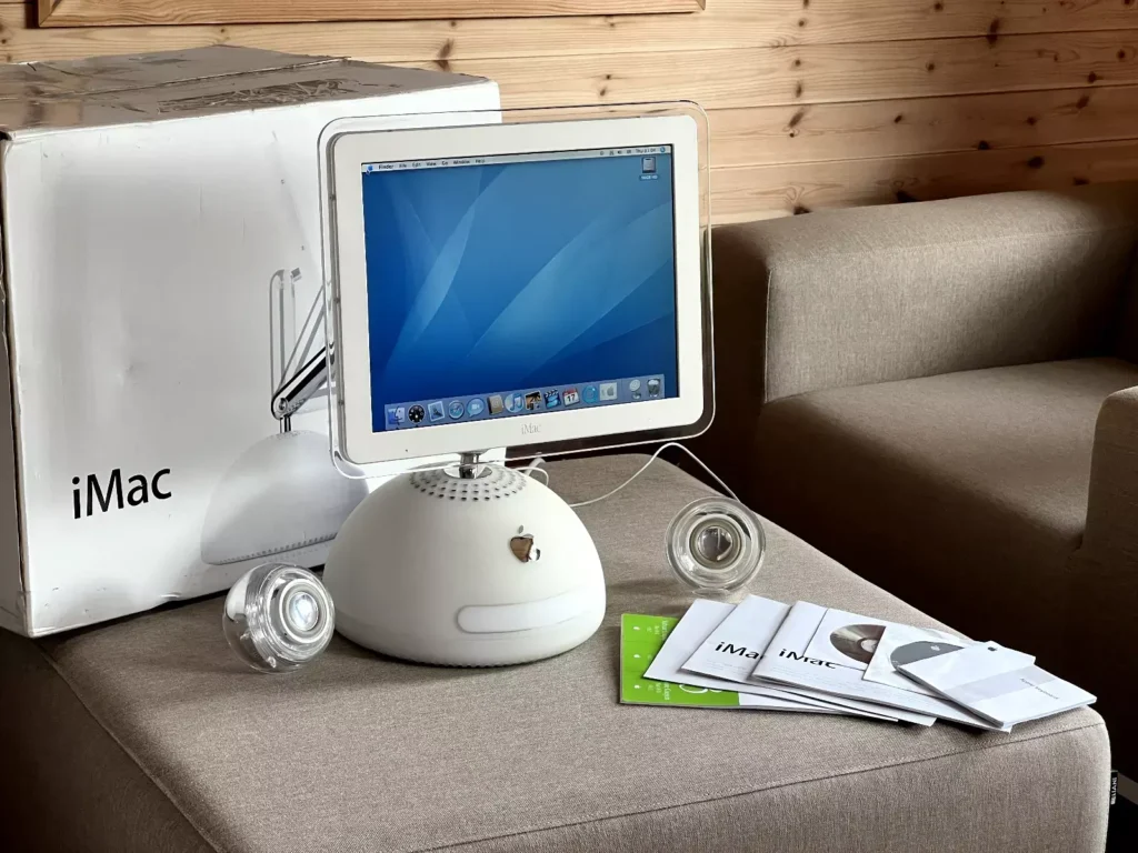 Apple iMac G4 s originální krabicí, klávesnicí, myší a manuály, vystavený na pohovce. Ikonický design s kulatou základnou a tenkým krkem podpírajícím monitor, typický pro model z roku 2003.