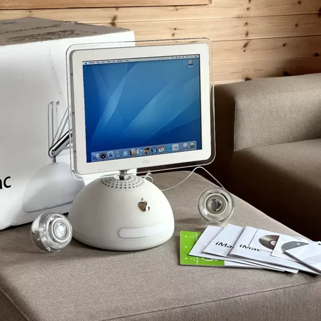 Apple iMac G4 s originální krabicí, klávesnicí, myší a manuály, vystavený na pohovce. Ikonický design s kulatou základnou a tenkým krkem podpírajícím monitor, typický pro model z roku 2003.