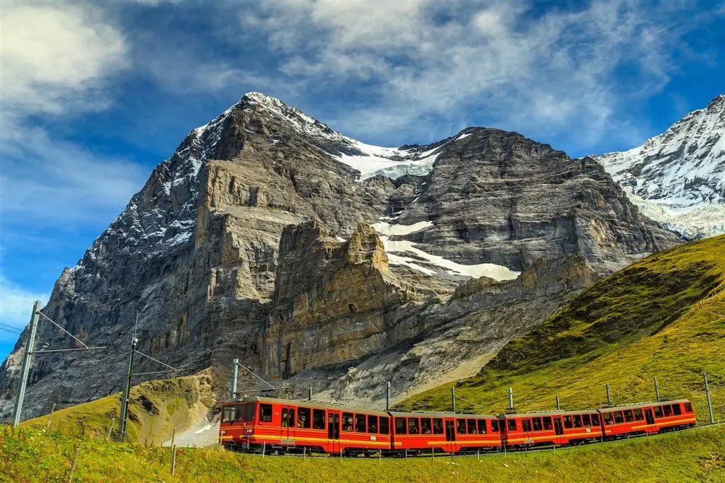 Červený vlak projíždějící pod majestátními švýcarskými horami na trase Glacier Express, s modrou oblohou a zasněženými vrcholky v pozadí