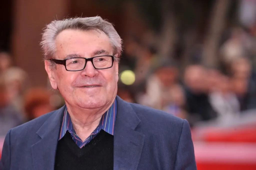 Miloš Forman s úsměvem na tváři při slavnostní premiéře, oblečený ve stylovém saku s košilí s barevným límcem.