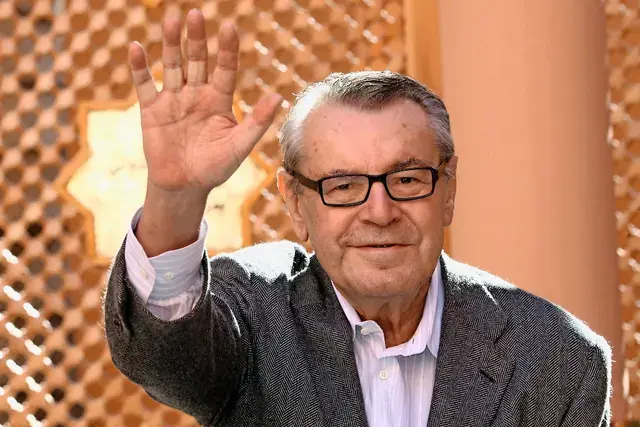  Miloš Forman mávající svým fanouškům s úsměvem na tváři, oblečený v elegantním saku, venku na veřejné akci.