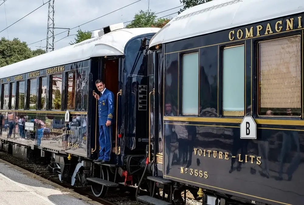 Průvodčí v modré uniformě stojící ve dveřích luxusního vlaku Orient Express, který je symbolem elegance a luxusu v železniční dopravě.