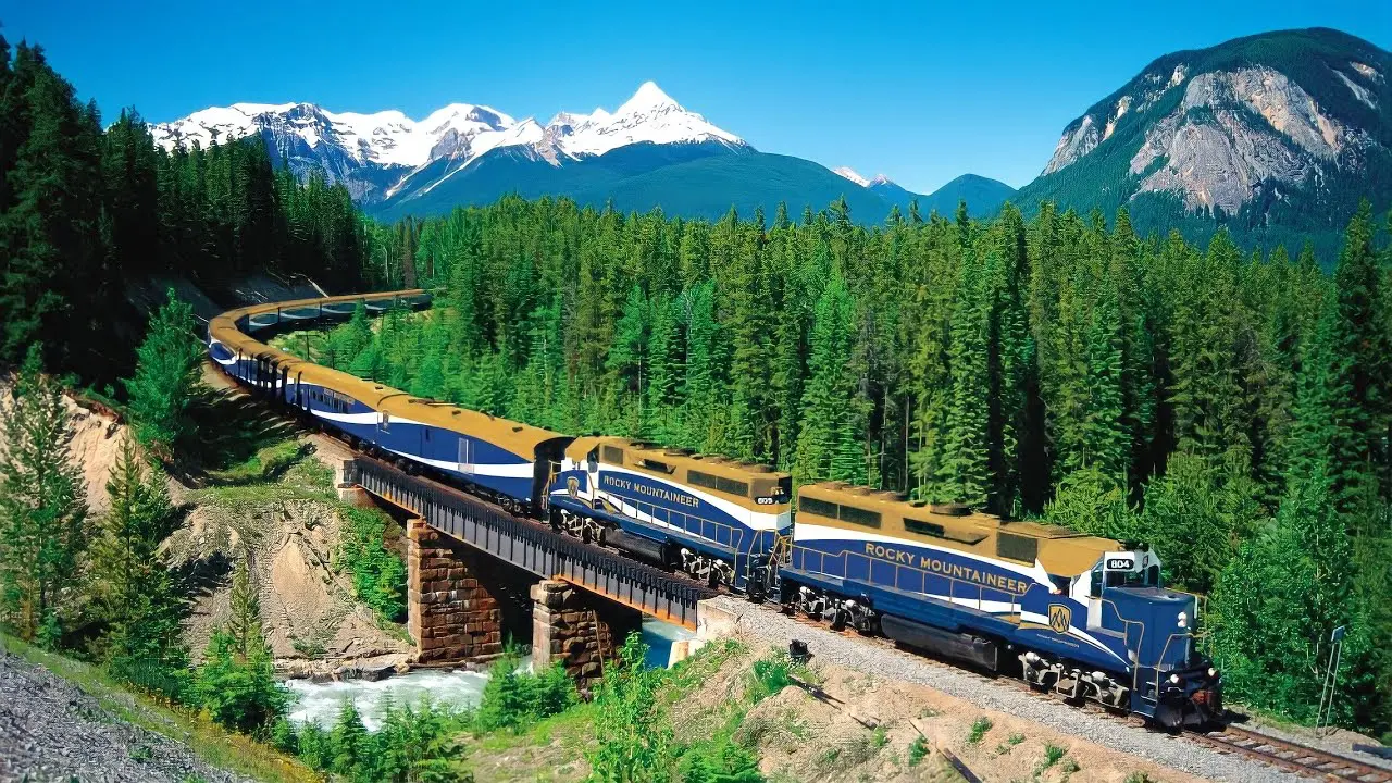 Vlak Rocky Mountaineer projíždějící zelenými lesy kanadských Skalnatých hor, s úchvatnými zasněženými vrcholky hor v pozadí.