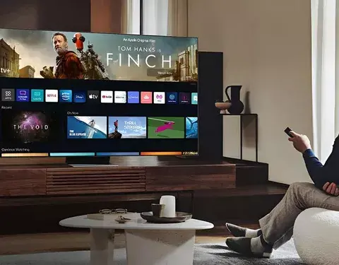 Osoba sedící v moderním obývacím pokoji, sledující chytrou televizi značky Samsung, na jejíž obrazovce je zobrazený film "Finch" s rozhraním aplikací jako Netflix, Apple TV a další.